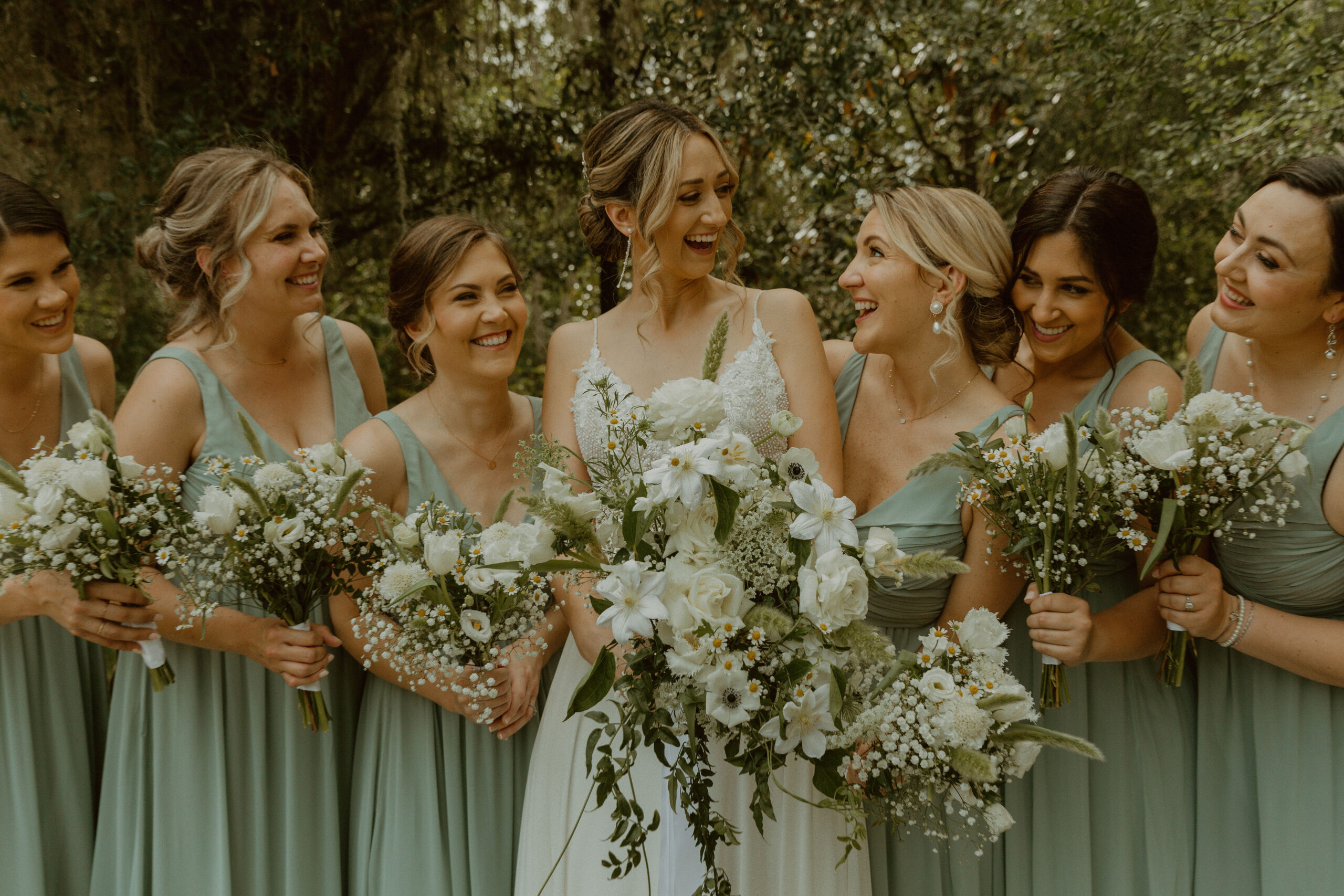 Radiant moment: Bride shares a photo, laughing with six bridesmaids, each in elegant green dresses and holding white flowers.