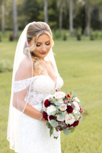 Timeless beauty: Bride holding a bouquet of red and white roses, looking down, adorned with perfect hair and makeup by Kristy's Artistry Design Team.