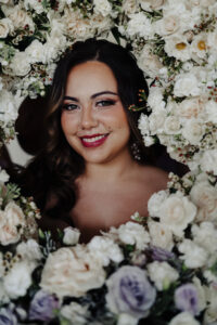 Captivating gaze: Bride, surrounded by a ring of white roses, looks directly at the camera, showcasing the artistry of hair and makeup by Kristy's Artistry Design Team.