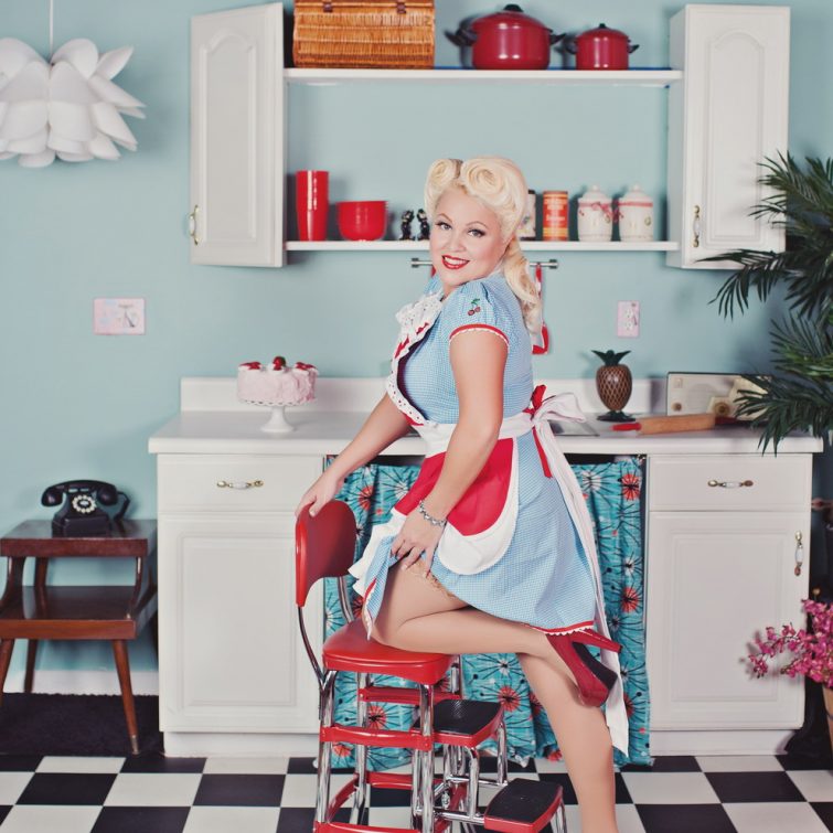 Blonde model in a blue dress with a red and white apron, posing with her knee on a chair in a kitchen setting. Hair and makeup by Kristy's Artistry Design Team for magazine photo spreads.