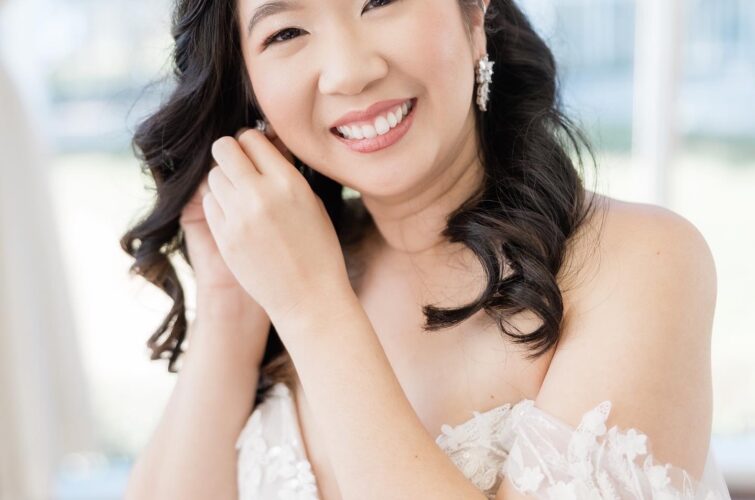 Smiling Asian bride wearing a tiara and putting on a right-side earring, showcasing beautiful hair and makeup by Kristy's Artistry Design Team (KADT)
