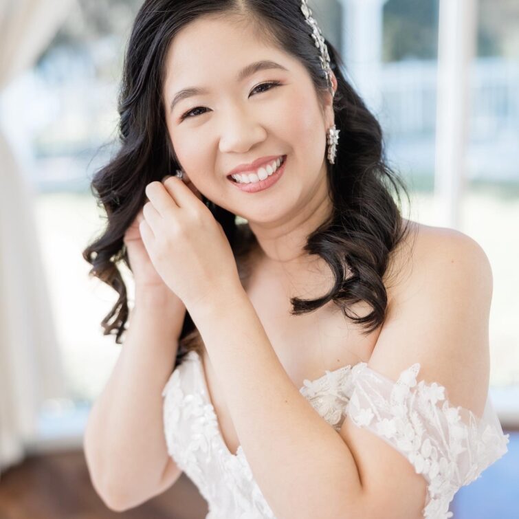 Smiling Asian bride wearing a tiara and putting on a right-side earring, showcasing beautiful hair and makeup by Kristy's Artistry Design Team (KADT)