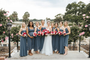 Joyful bride and bridesmaids, adorned with flawless hair and makeup by Kristy's Artistry Design Team, pose against a scenic backdrop of a water fountain and sky, holding vibrant flowers.