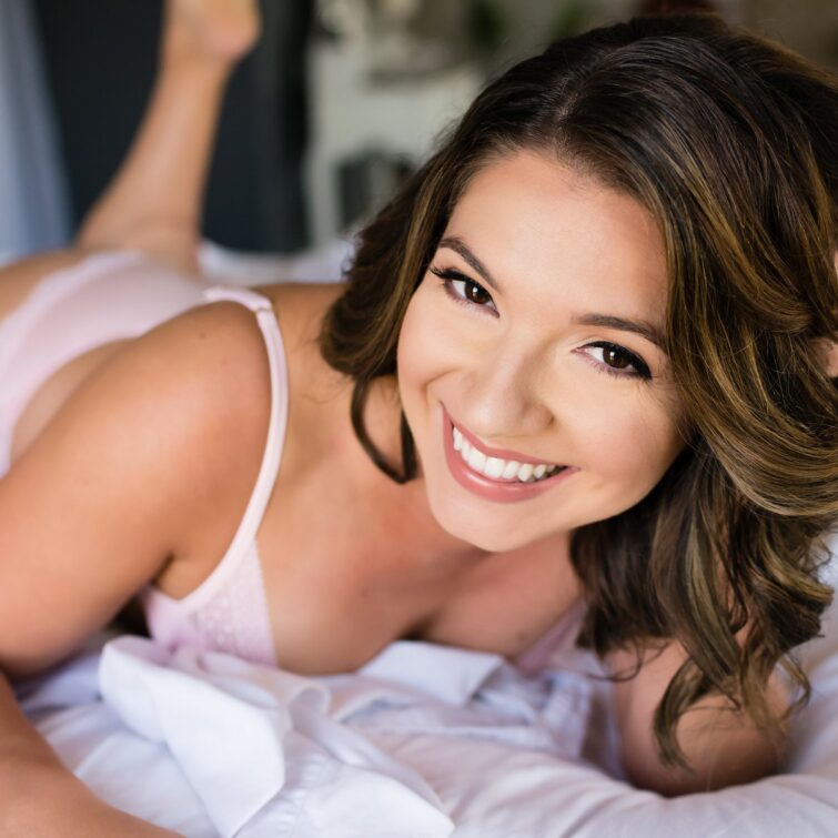 Image of a girl smiling for her boudoir photo, lying on a bed in the studio, with her hair and makeup beautifully done by Kristy's Artistry Design Team.