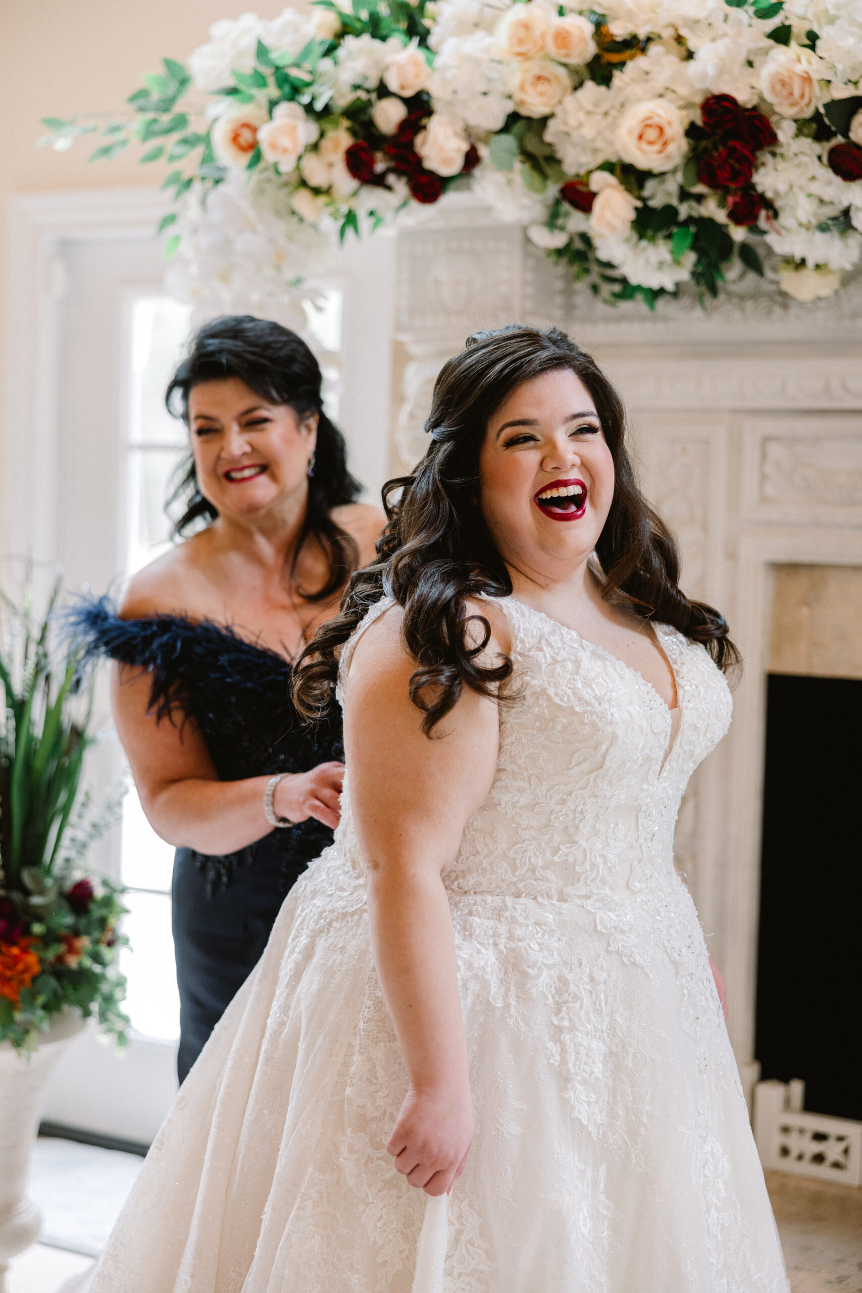 other of the bride zipping up the bride's dress, both smiling and laughing with hair and makeup done by Kristys Artistry Design Team.