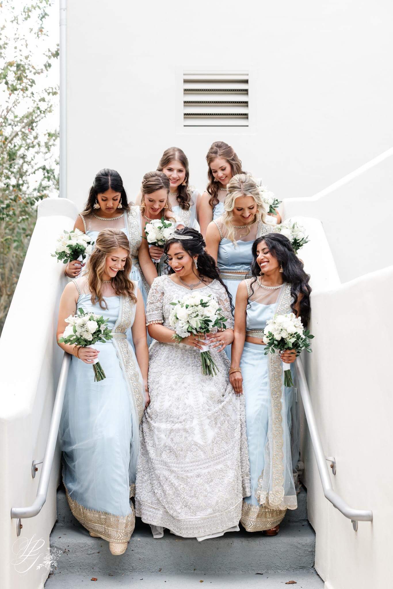 Bride and bridal party walking down the stairs of the venue with hair and makeup by Kristy's Artistry Design Team, all holding flowers, bride wearing a tiara.