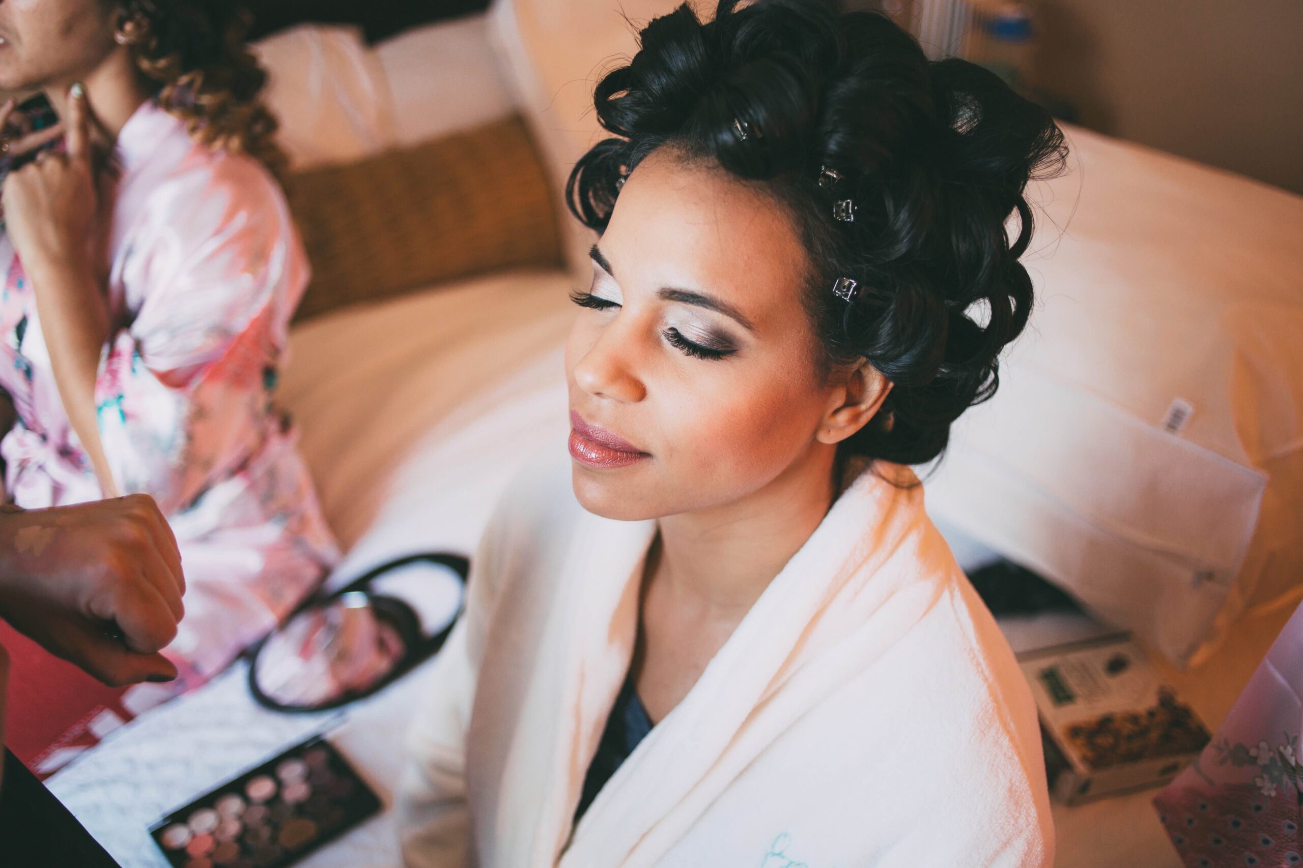 Bride smiling while getting her makeup done on her hotel bed next to her bridesmaid, with Kristy's Artistry Design Team providing mobile hair and makeup services for the Orlando wedding.