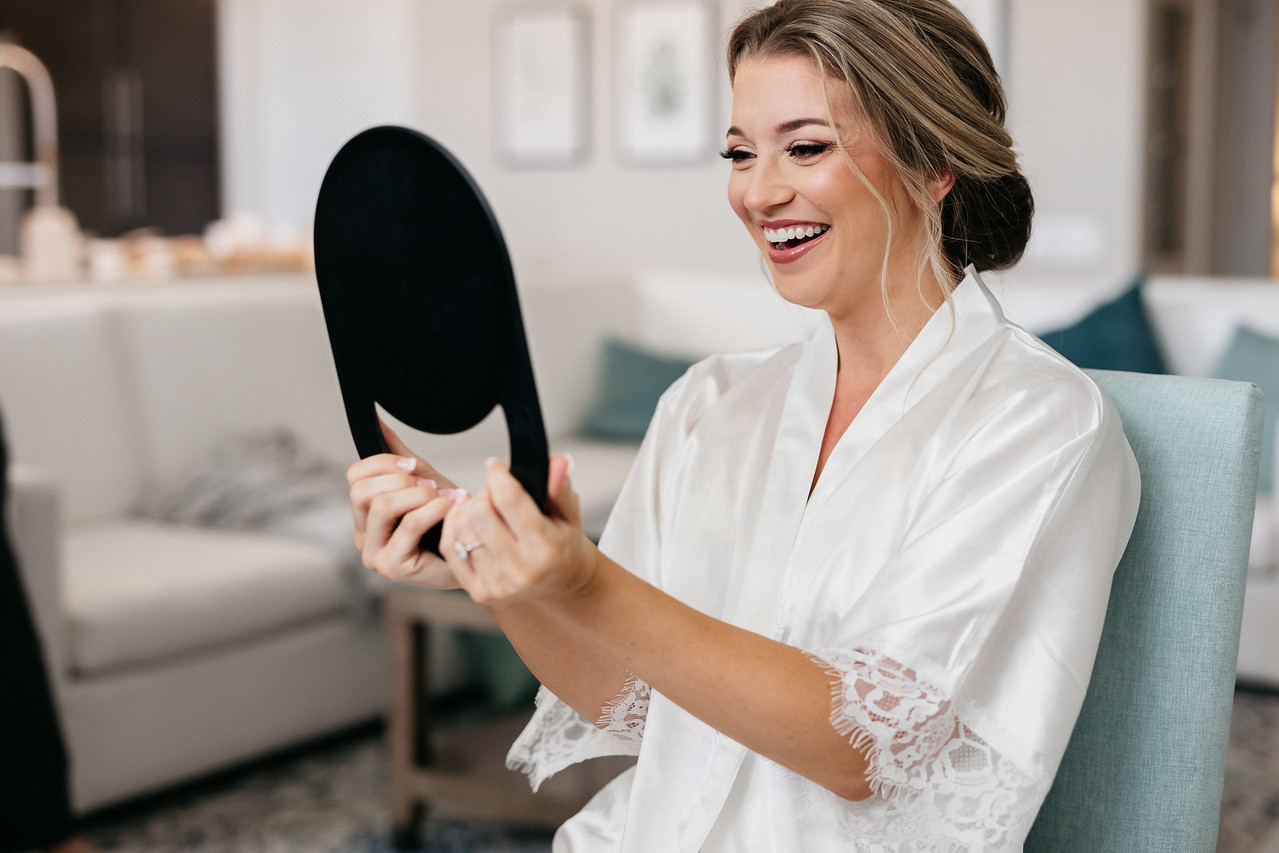 Bride holding a mirror and smiling at her final makeup, showcasing why brides trust Kristy's Artistry Design Team.