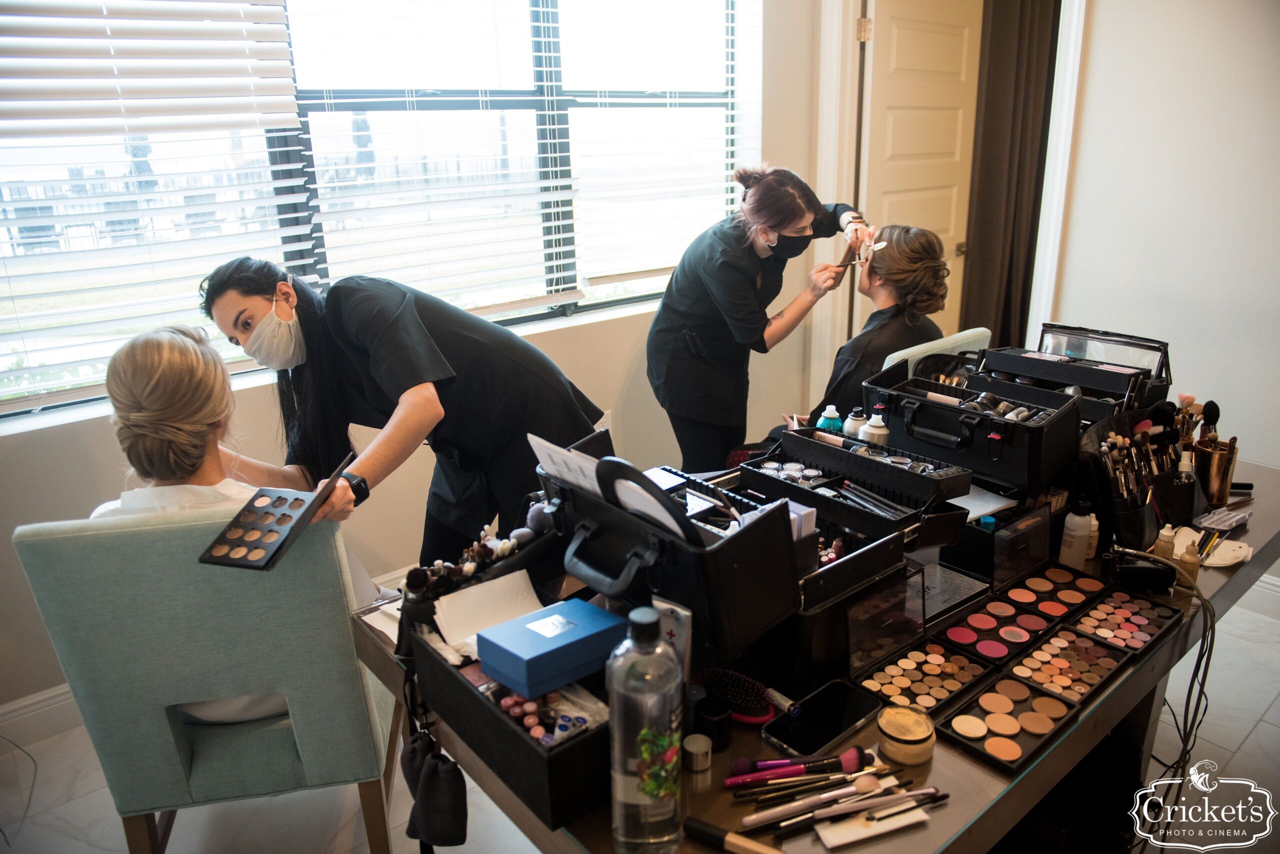 Two artists working side by side with makeup kits open on a table, applying makeup to a bride and bridesmaid, both with updos by Kristy's Artistry Design Team in Orlando.