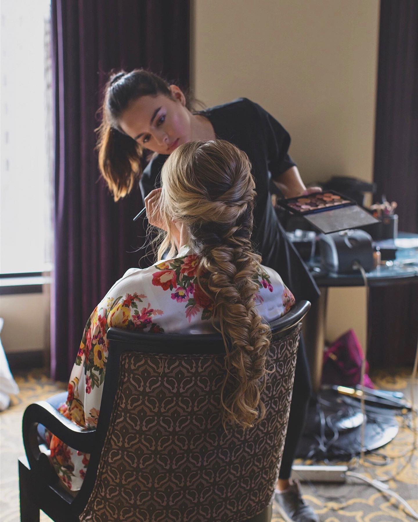 Makeup artist applying makeup to a bride with a brush in one hand and a color palette in the other, showcasing the bride's intricate down do hairstyle by Kristy's Artistry Design Team.