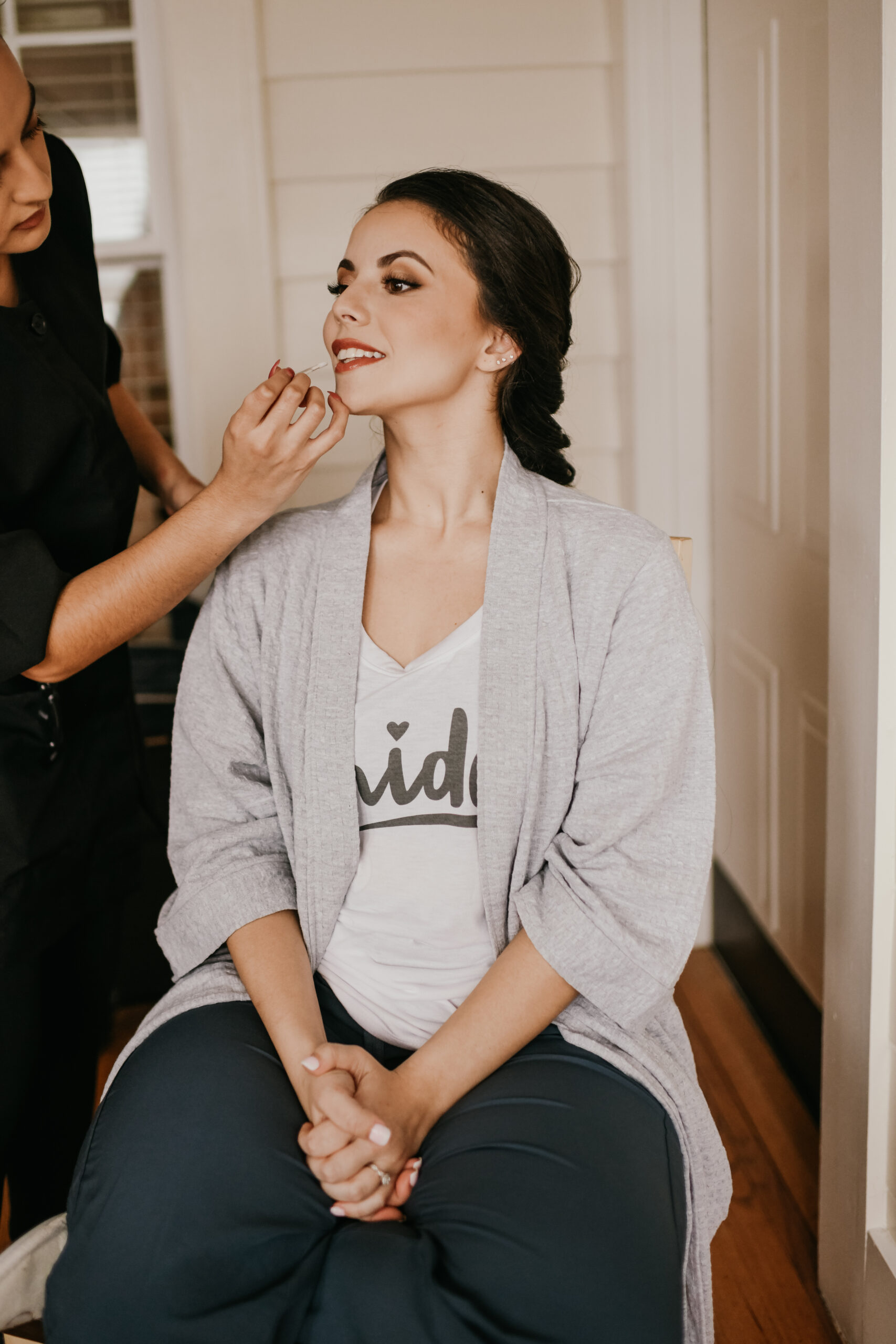 Smiling bride sitting pre-wedding while a makeup artist from Kristy's Artistry Design Team applies finishing touches to her lips.