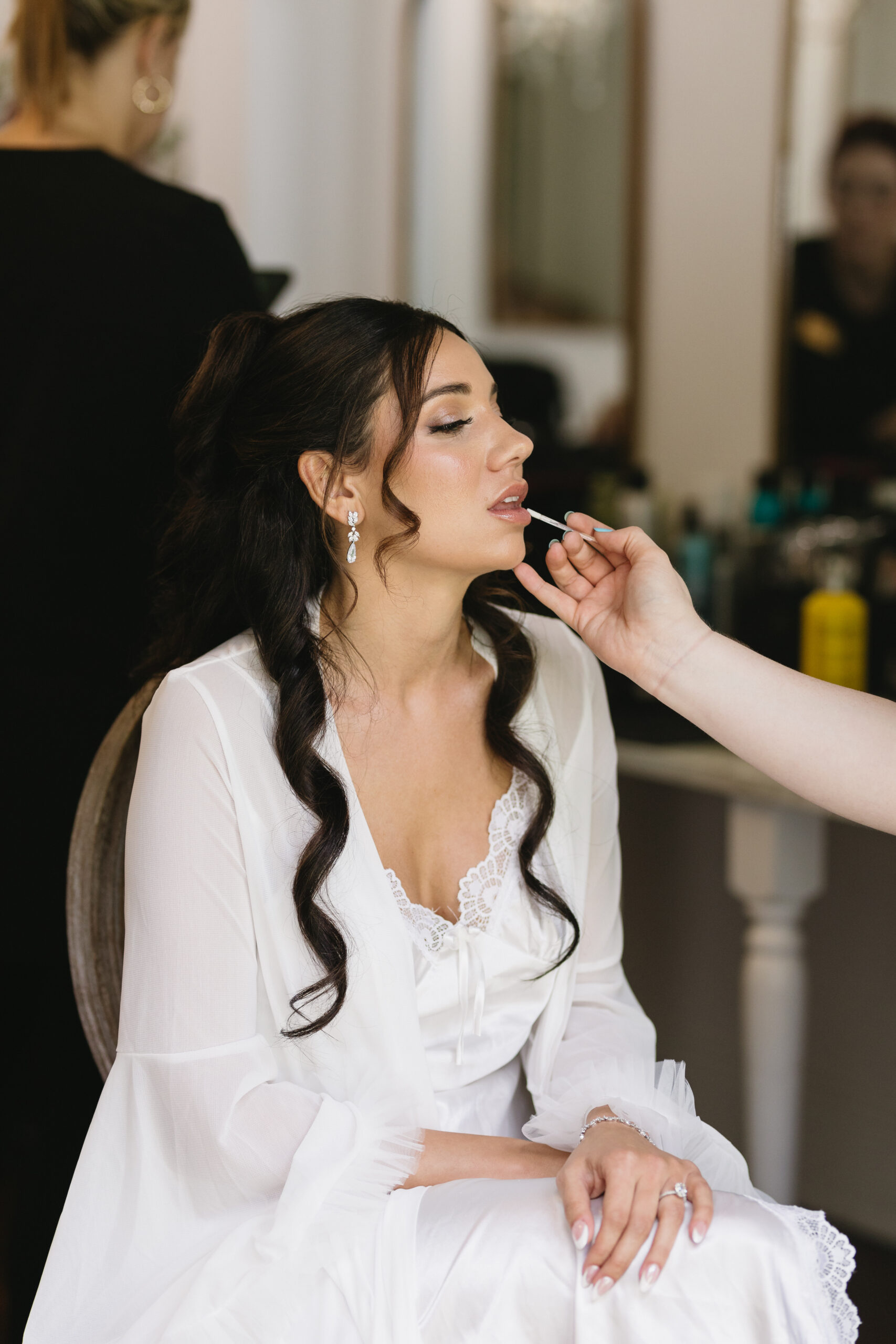 Bride receiving finishing touches on her lip balm while bridesmaids have their hair and makeup done in the background.